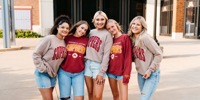 5 girls standing next to each other smiling wearing and wearing University of Minnesota apparel in front of the football stadium gates