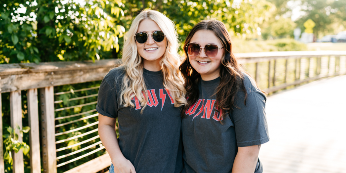 two girls on a bridge with trees in the background wearing the same faded gray t-shirt and sunglasses