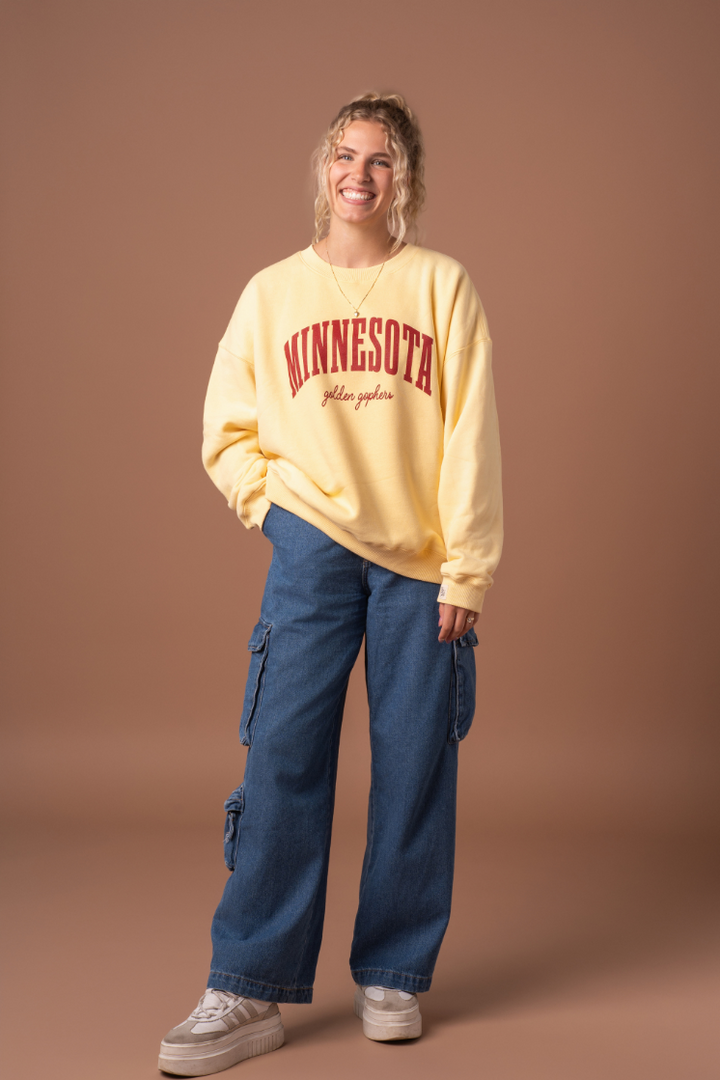 full body image of a girl wearing jeans and a butter yellow crewneck with Minnesota Golden Gophers screenprinted in maroon across the chest
