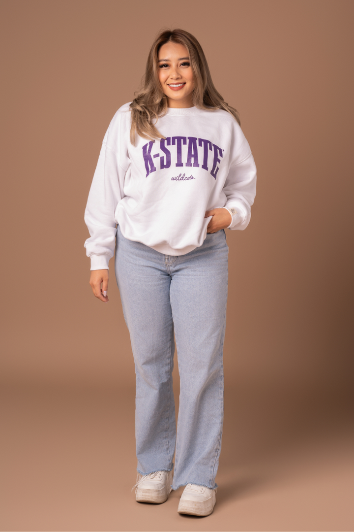 A full body photo of a girl wearing an oversized fleece lined white crew neck with a large purple K-State Wildcats design on the front.