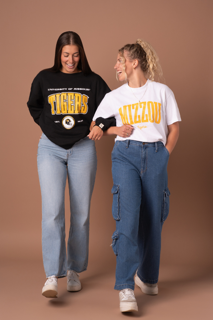 A photo of two girls linking arms. They are wearing a black crew neck with a large University of Missouri Tigers design on the front and a white tucked in t-shirt with a Mizzou Tigers design in gold.