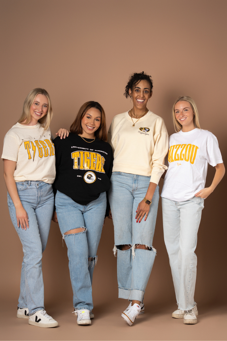 A photo of four girls wearing University of Missouri tops including a t-shirt, crew neck, and Supersoft crew neck. They all have different Mizzou Tigers designs on the front of the top. They are wearing these with jeans and sneakers.