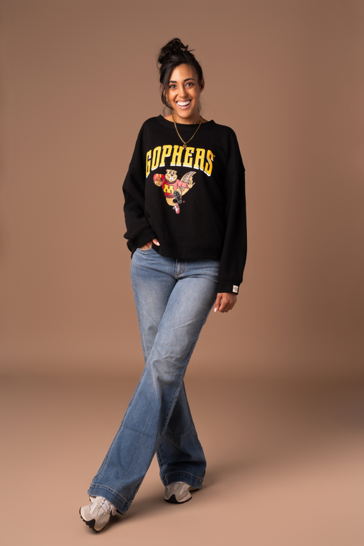 full body image of girl wearing jeans and a black crewneck with Gophers screenprinted in gold across the chest with a Goldy graphic holding a football in maroon and gold decoration