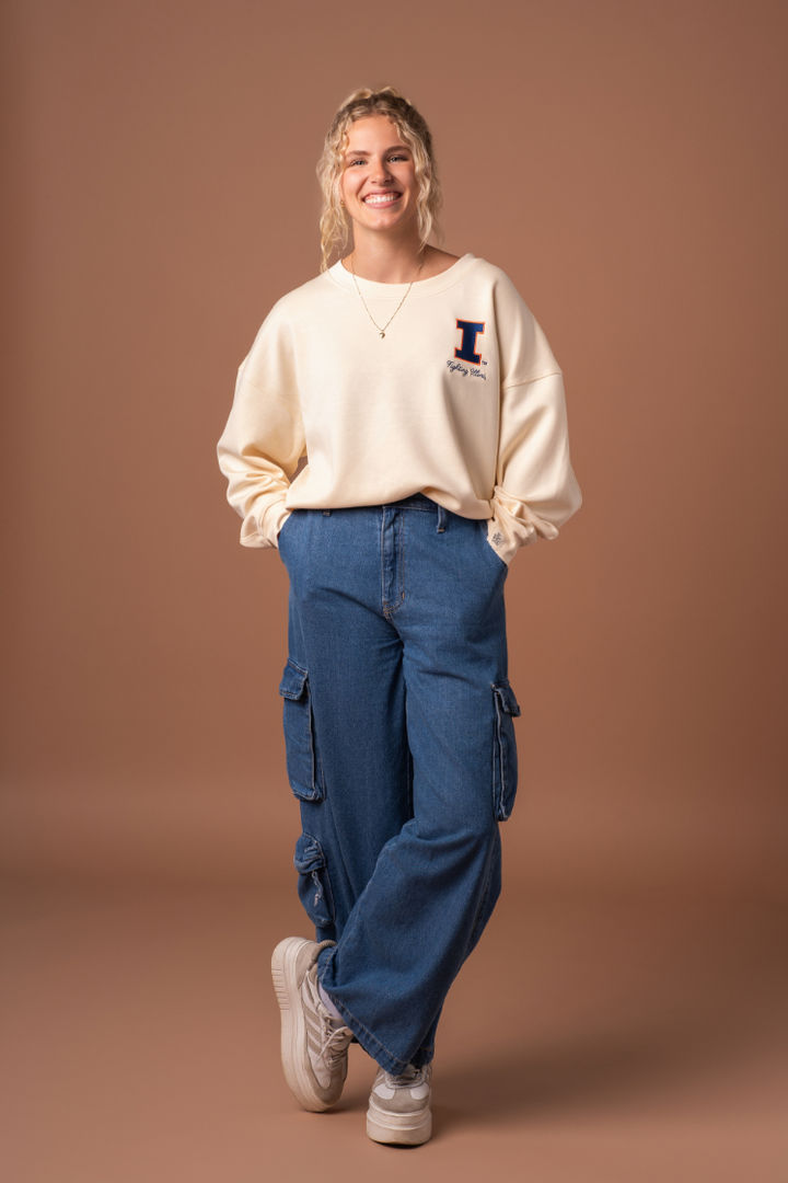 A woman with her hands in her pockets wearing a very soft, athleisure type ivory crew neck sweatshirt with an embroidered University of Illinois logo and cursive "fighting Illini" text below it.