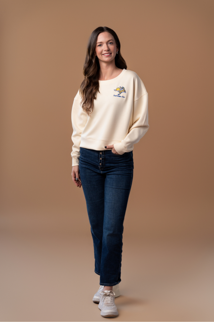 a full body image of a girl wearing jeans with a bone colored waist-length crewneck with an embroidered SDSU Jackrabbits logo on the left pocketside