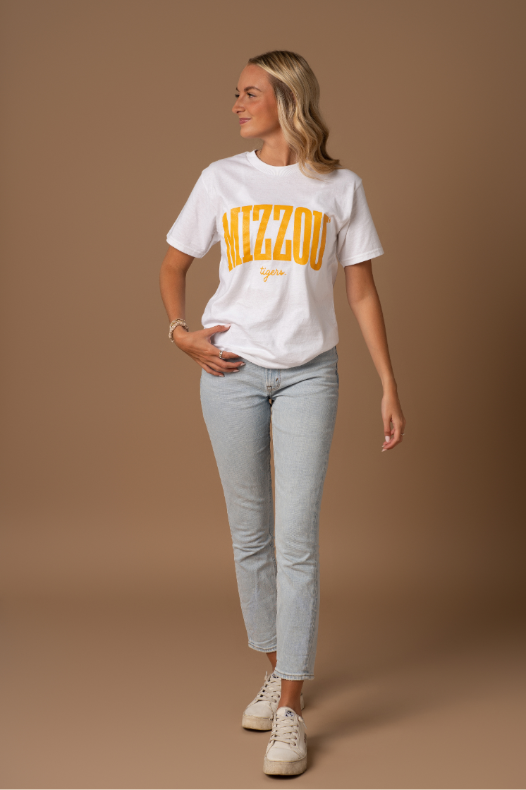 A full body photo of a girl wearing a white t-shirt with a Mizzou Tigers design on it in gold. She is wearing this with blue jeans and sneakers.