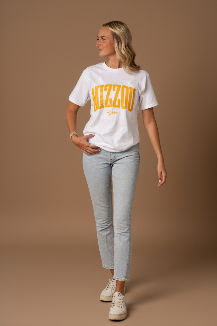 A full body photo of a girl wearing a white t-shirt with a Mizzou Tigers design on it in gold. She is wearing this with blue jeans and sneakers.