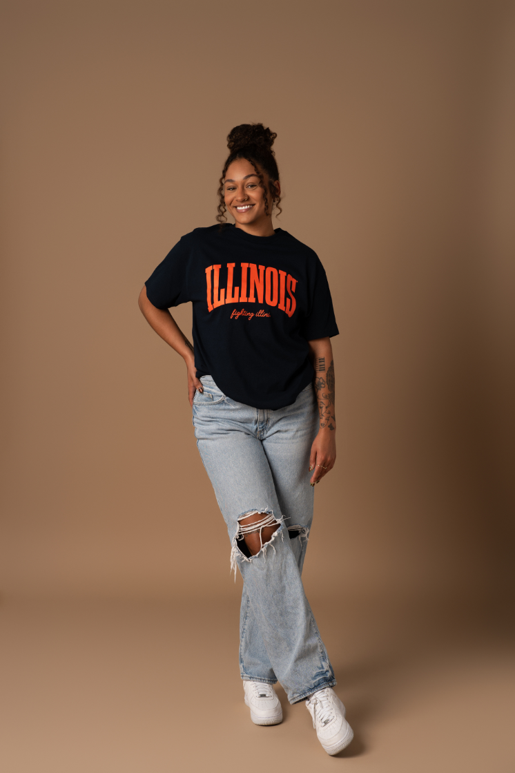 A model with her hand on her hip wearing jeans a navy blue comfortable and understated University of Illinois women's t-shirt that has team colors orange "Illinois Fighting Illini" screenprinted decoration.