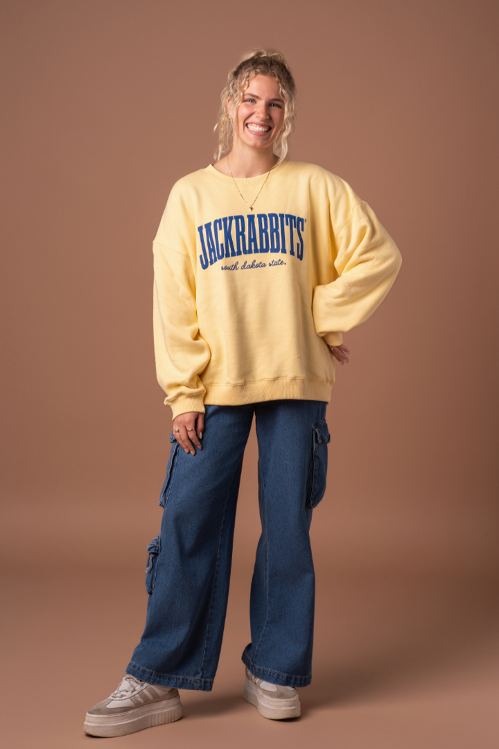 a full body image of a girl smiling, wearing jeans and a yellow crewneck with Jackrabbits, south dakota state screenprinted in blue