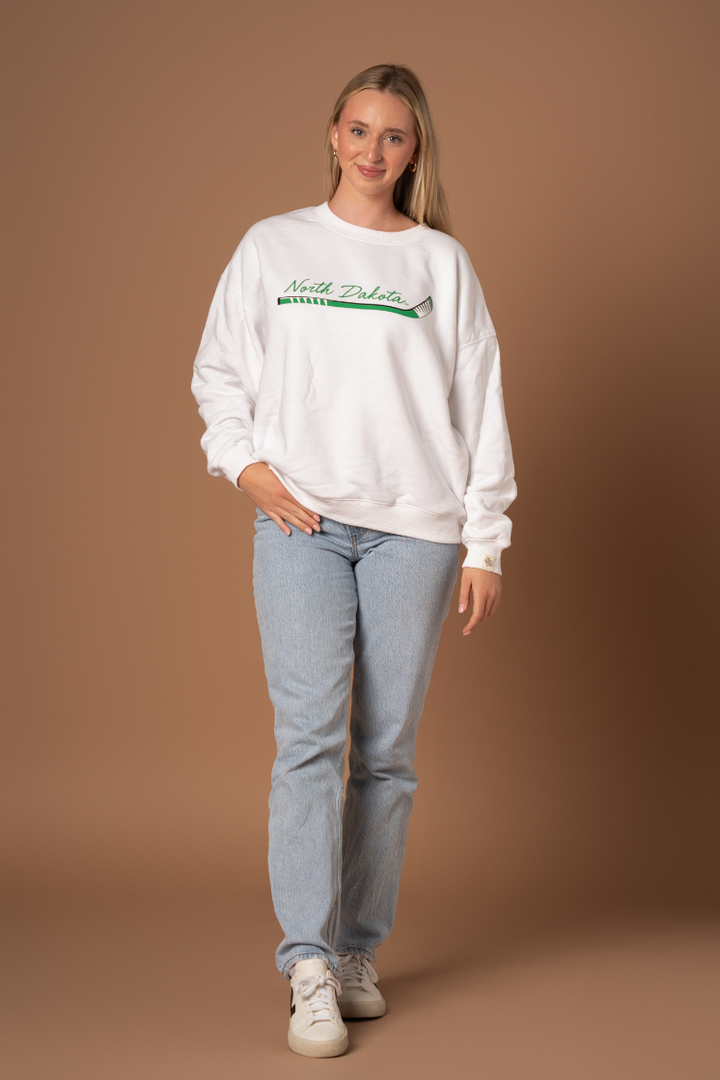 a full body image of a girl wearing jeans and white crewneck that says North Dakota in green with a hockey stick graphic below it
