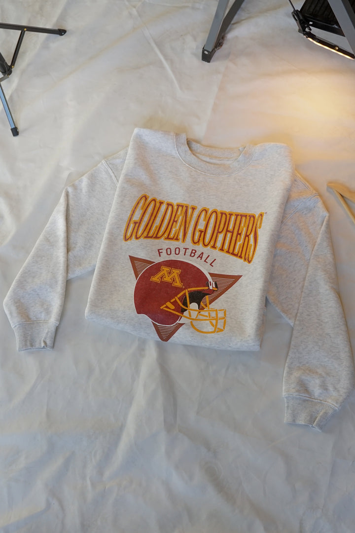 flat lay image of an ash gray crewneck with Golden Gophers Football screenprinted with a distressed football graphic in maroon and gold