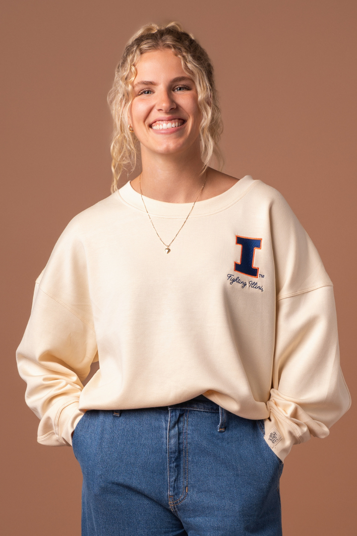 Close up of a woman wearing a very soft, athleisure type ivory crew neck sweatshirt with an embroidered University of Illinois logo and cursive "fighting Illini" text below it.