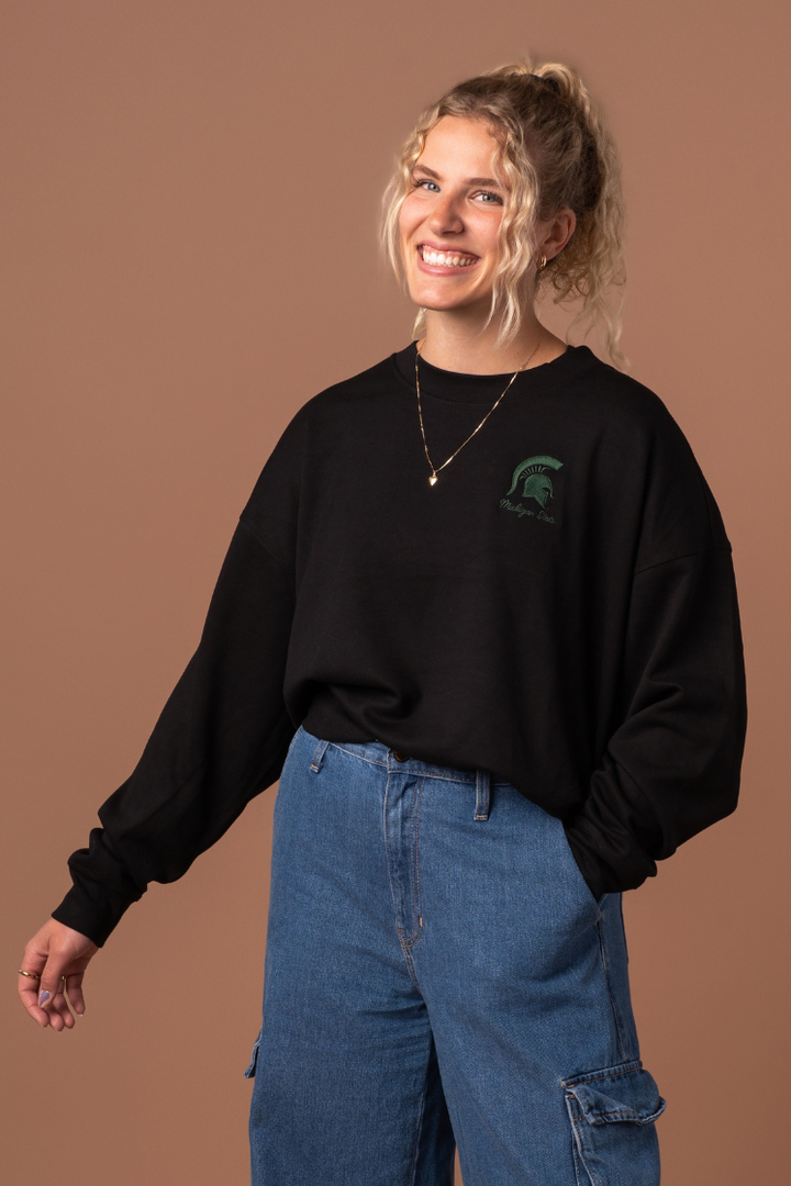 close up picture of a girl wearing a black Michigan State Supersoft Waist Length Crew Neck with a green embroidered  Spartan logo