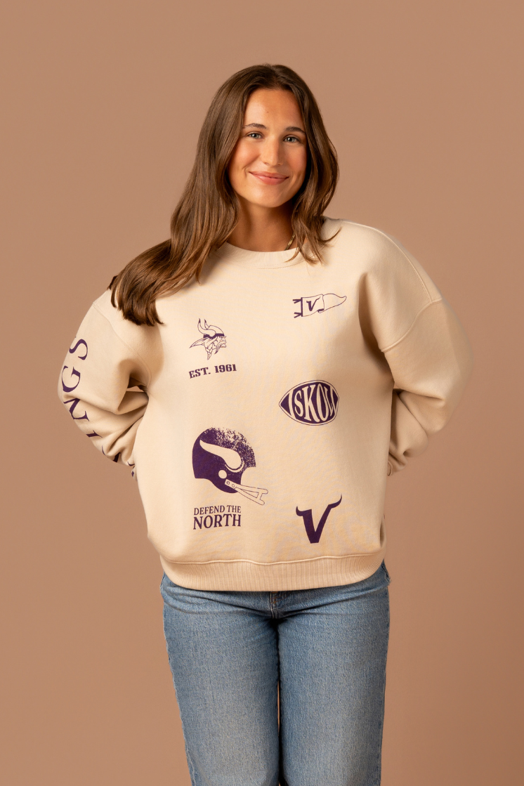 Woman wearing a tan crew neck sweatshirt with five, small purple Minnesota Vikings graphics scattered around the front with purple 'Vikings' text down the arm