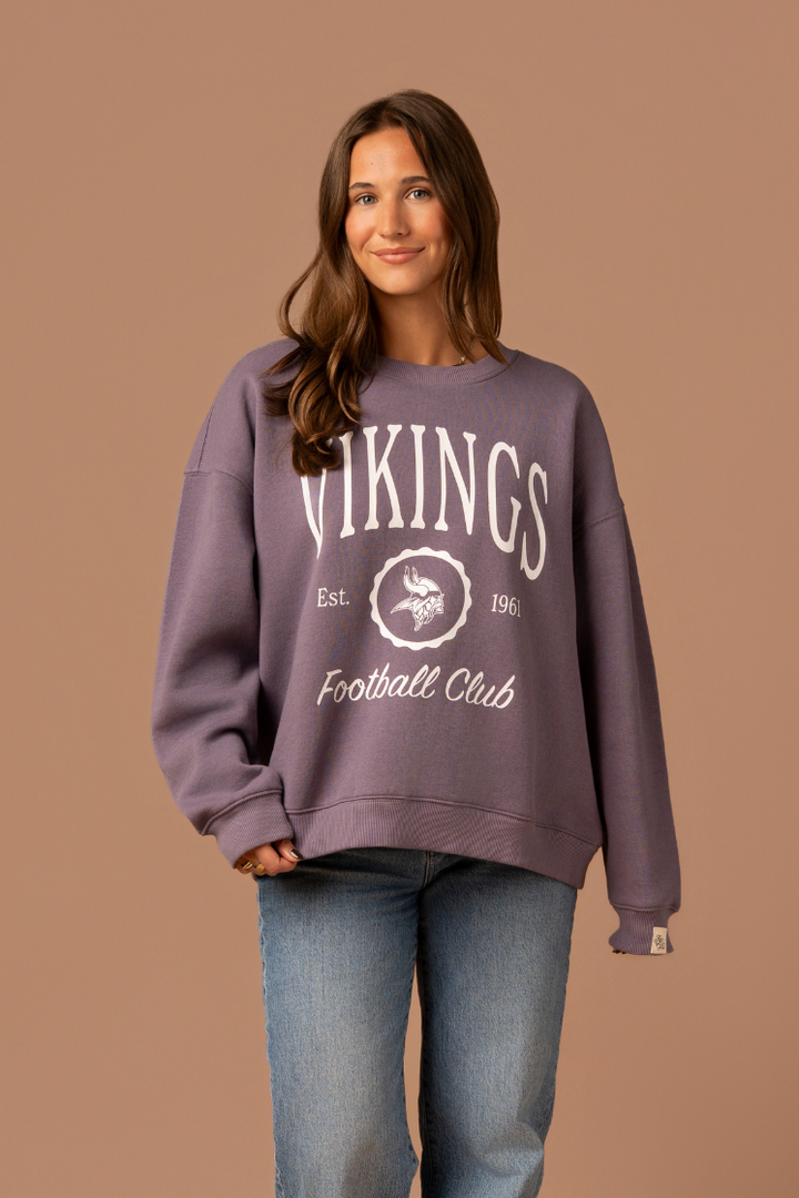 Woman wearing a purple crew neck sweatshirt with white 'Vikings Football Club' and 'EST 1961' text and a white Vikings logo in the center