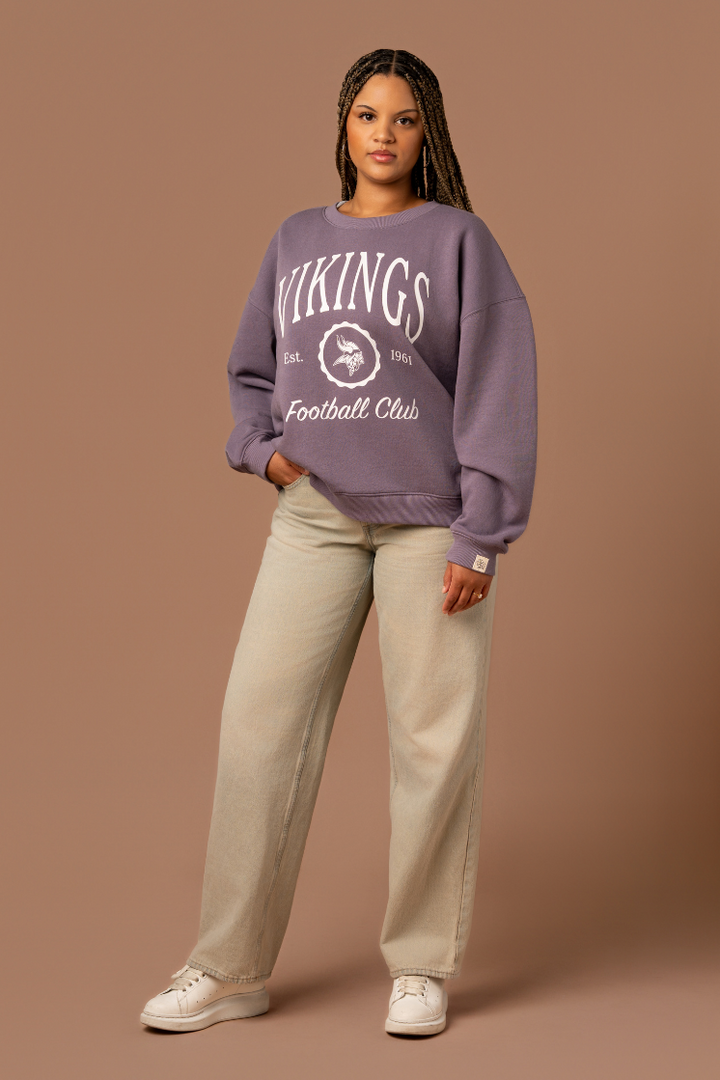 Woman wearing a purple crew neck sweatshirt with white 'Vikings Football Club' and 'EST 1961' text and a white Vikings logo in the center