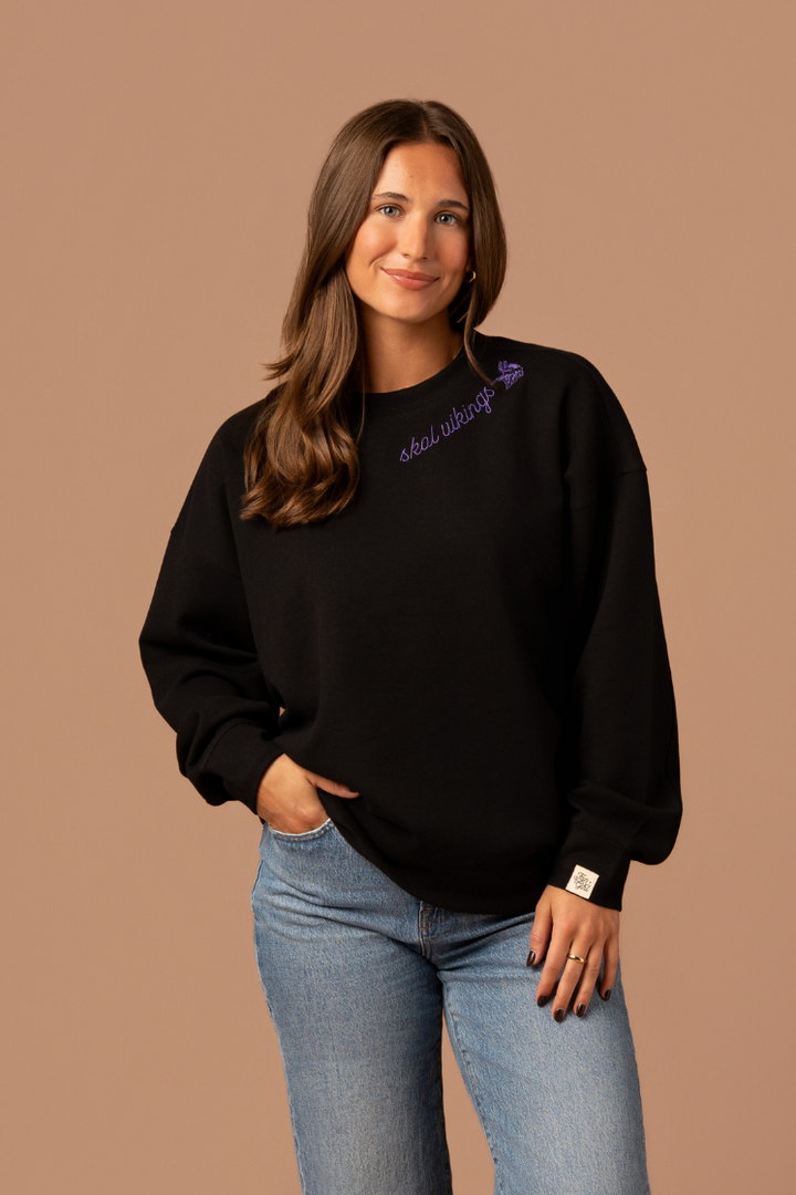 Woman wearing a black crew neck sweatshirt with purple cursive 'Skol Vikings' text and a logo Vikings logo along the neckline