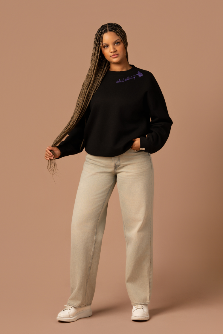 Woman wearing a black crew neck sweatshirt with purple cursive 'Skol Vikings' text and a logo Vikings logo along the neckline