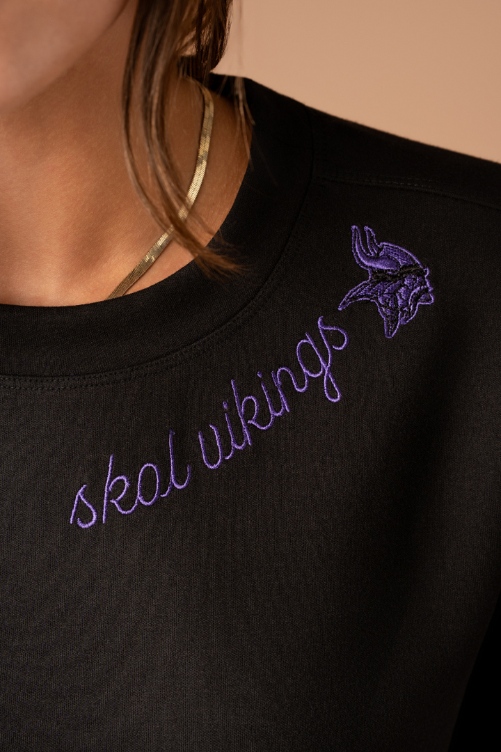 A black crew neck sweatshirt with purple cursive 'Skol Vikings' text and a logo Vikings logo along the neckline