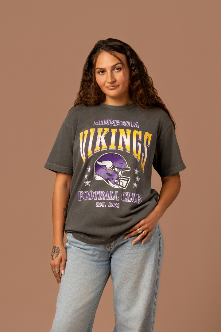 Woman wearing a faded black t-shirt with gold and purple 'Minnesota Vikings Football Club' and 'EST. 1961' text with a purple Vikings helmet graphic in the center
