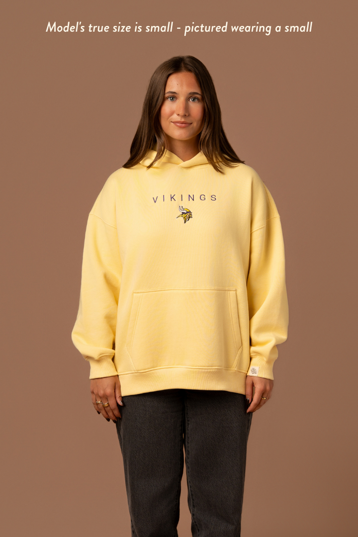 Woman wearing a yellow hoodie with purple 'Vikings' text and a small Vikings logo in the center. Caption reads "model's true size is small- pictured wearing a small"