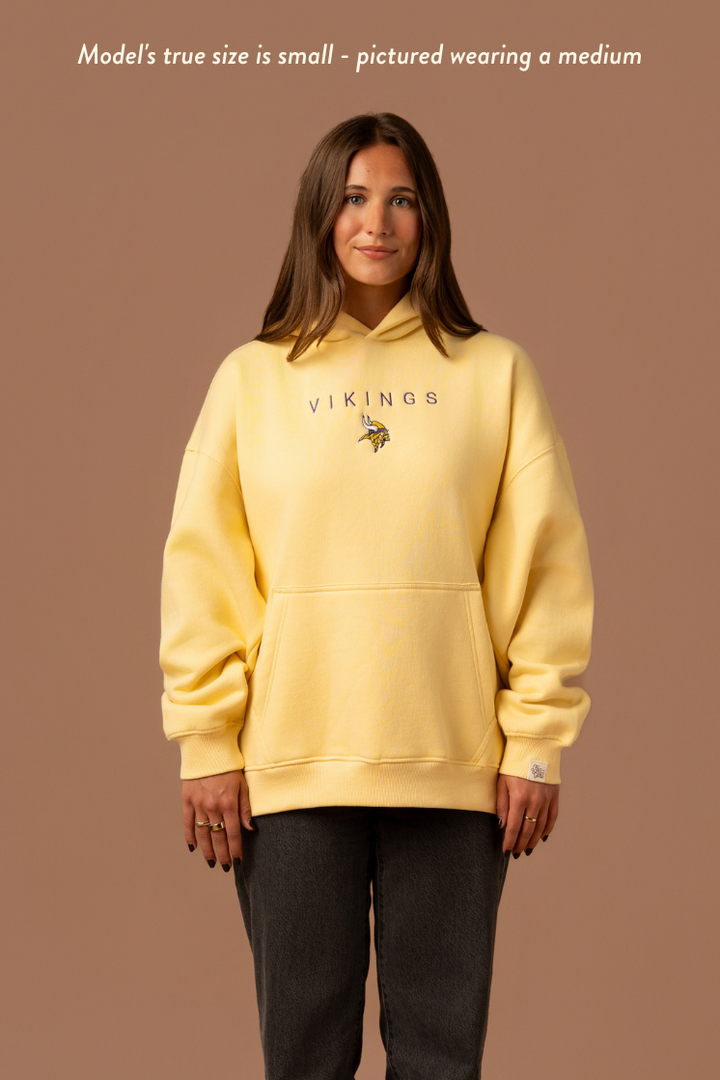Woman wearing a yellow hoodie with purple 'Vikings' text and a small Vikings logo in the center. Caption reads "model's true size is small- pictured wearing a medium"