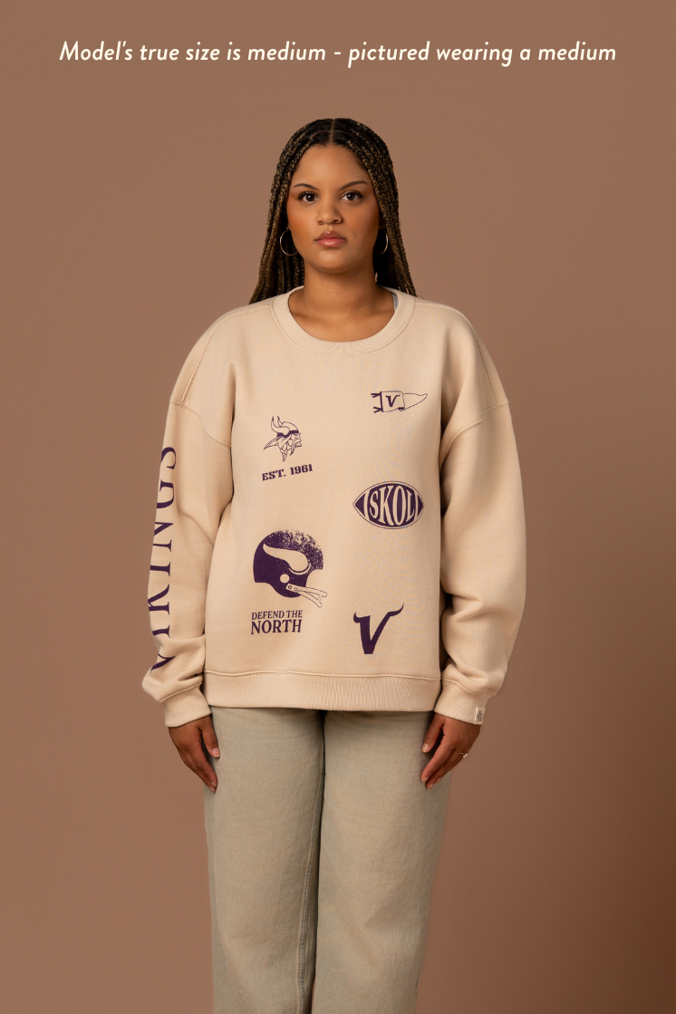 Woman wearing a tan crew neck sweatshirt with five, small purple Minnesota Vikings graphics scattered around the front with purple 'Vikings' text down the arm. Caption reads "model's true size is medium- pictures wearing a medium"