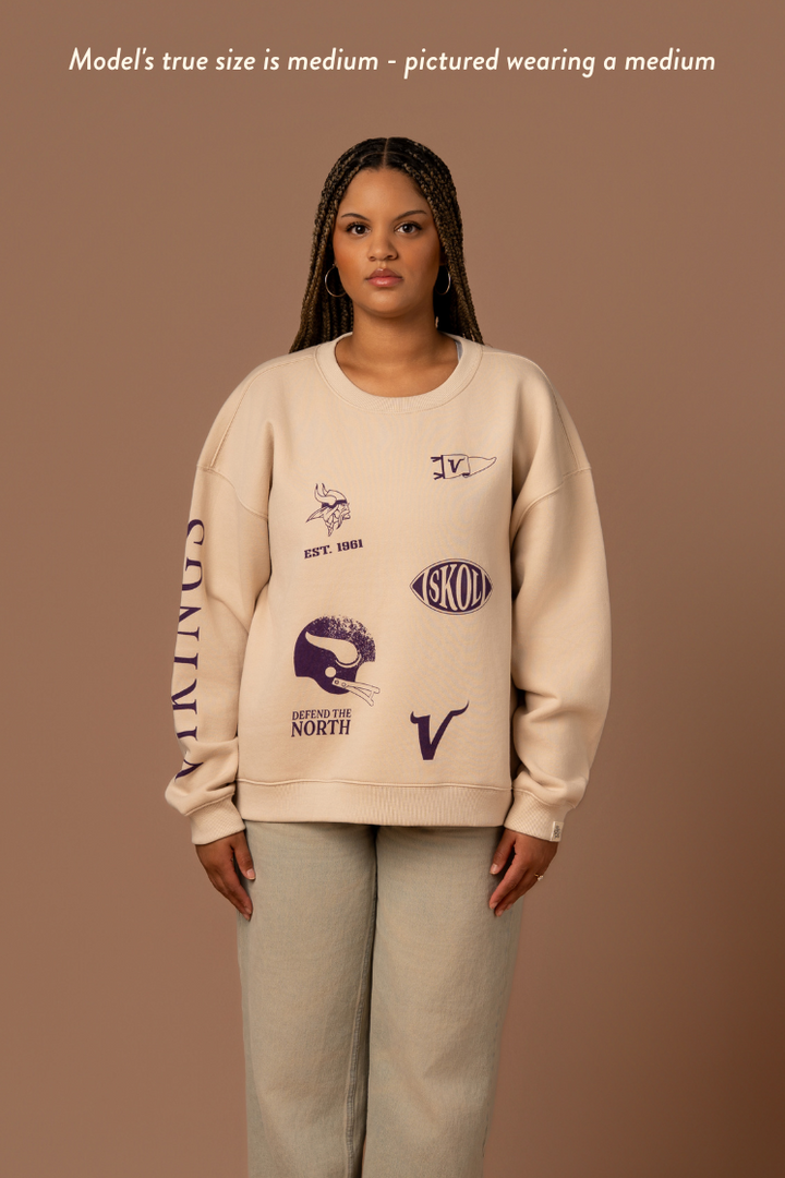 Woman wearing a tan crew neck sweatshirt with five, small purple Minnesota Vikings graphics scattered around the front with purple 'Vikings' text down the arm. Caption reads "model's true size is medium- pictures wearing a medium"
