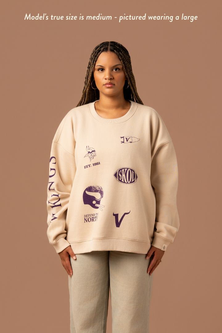 Woman wearing a tan crew neck sweatshirt with five, small purple Minnesota Vikings graphics scattered around the front with purple 'Vikings' text down the arm. Caption reads "model's true size is medium- pictures wearing a large"