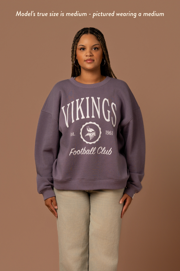 Woman wearing a purple crew neck sweatshirt with white 'Vikings Football Club' and 'EST 1961' text and a white Vikings logo in the center. Caption reads "Model's true size is medium - pictured wearing a medium"