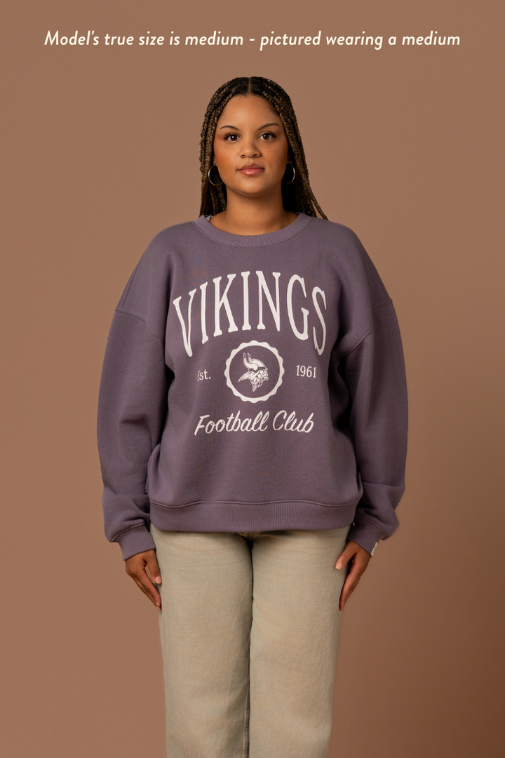 Woman wearing a purple crew neck sweatshirt with white 'Vikings Football Club' and 'EST 1961' text and a white Vikings logo in the center. Caption reads "Model's true size is medium - pictured wearing a medium"