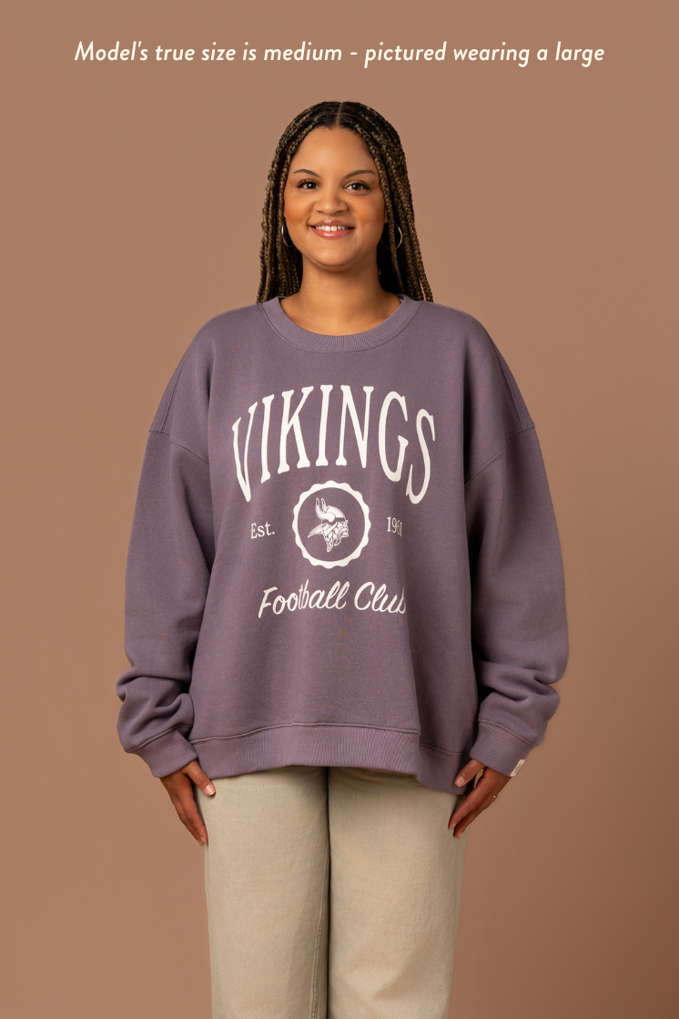 Woman wearing a purple crew neck sweatshirt with white 'Vikings Football Club' and 'EST 1961' text and a white Vikings logo in the center. Caption reads "Model's true size is medium - pictured wearing a large"
