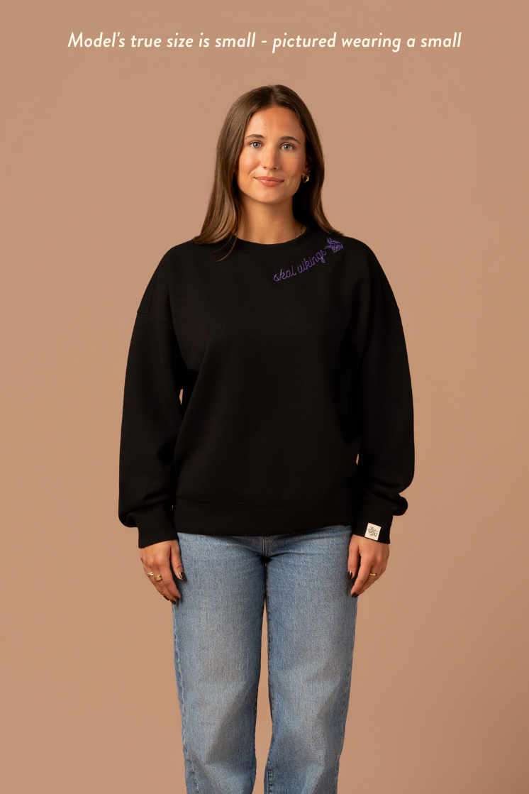 Woman wearing a black crew neck sweatshirt with purple cursive 'Skol Vikings' text and a logo Vikings logo along the neckline. Caption reads "model's true size is small- pictured wearing a small"