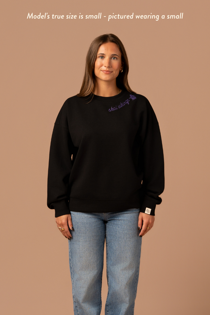 Woman wearing a black crew neck sweatshirt with purple cursive 'Skol Vikings' text and a logo Vikings logo along the neckline. Caption reads "model's true size is small- pictured wearing a small"