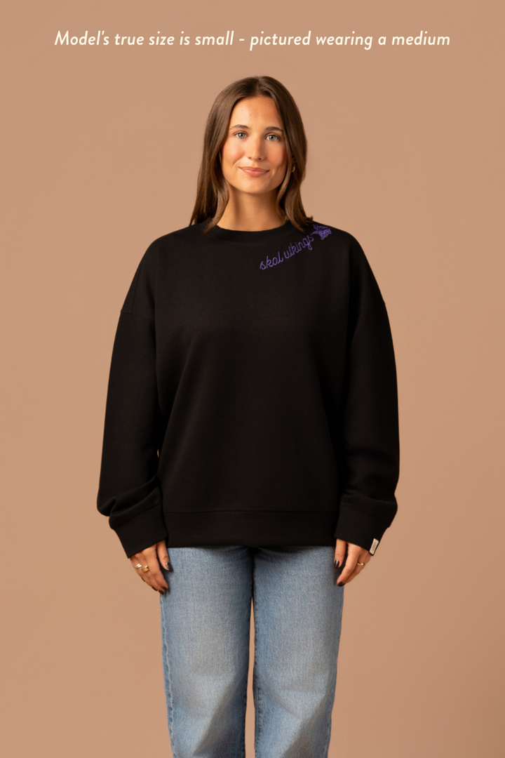 Woman wearing a black crew neck sweatshirt with purple cursive 'Skol Vikings' text and a logo Vikings logo along the neckline. Caption reads "model's true size is small- pictured wearing a medium"