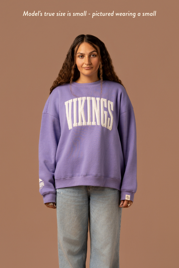 Woman wearing a purple crew neck sweatshirt with large, cream 'Vikings' text. Caption reads "model's true size is small- pictured wearing a small"
