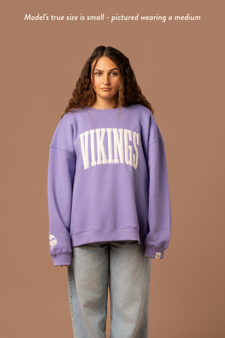 Woman wearing a purple crew neck sweatshirt with large, cream 'Vikings' text. Caption reads "model's true size is small- pictured wearing a medium"