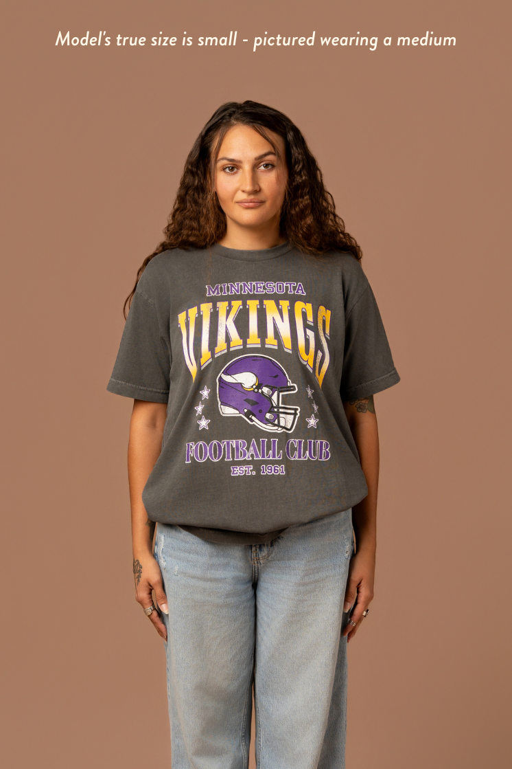 Woman wearing a faded black t-shirt with gold and purple 'Minnesota Vikings Football Club' and 'EST. 1961' text with a purple Vikings helmet graphic in the center. Caption reads "model's true size is small - pictured wearing a medium"