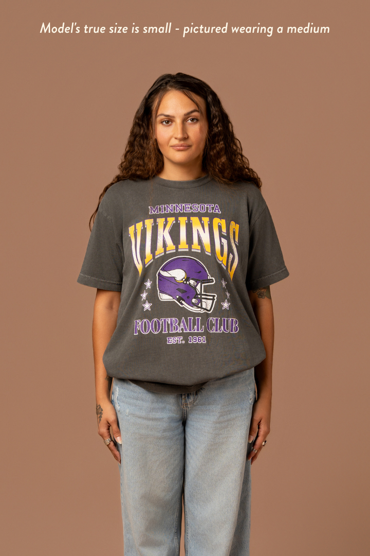 Woman wearing a faded black t-shirt with gold and purple 'Minnesota Vikings Football Club' and 'EST. 1961' text with a purple Vikings helmet graphic in the center. Caption reads "model's true size is small - pictured wearing a medium"