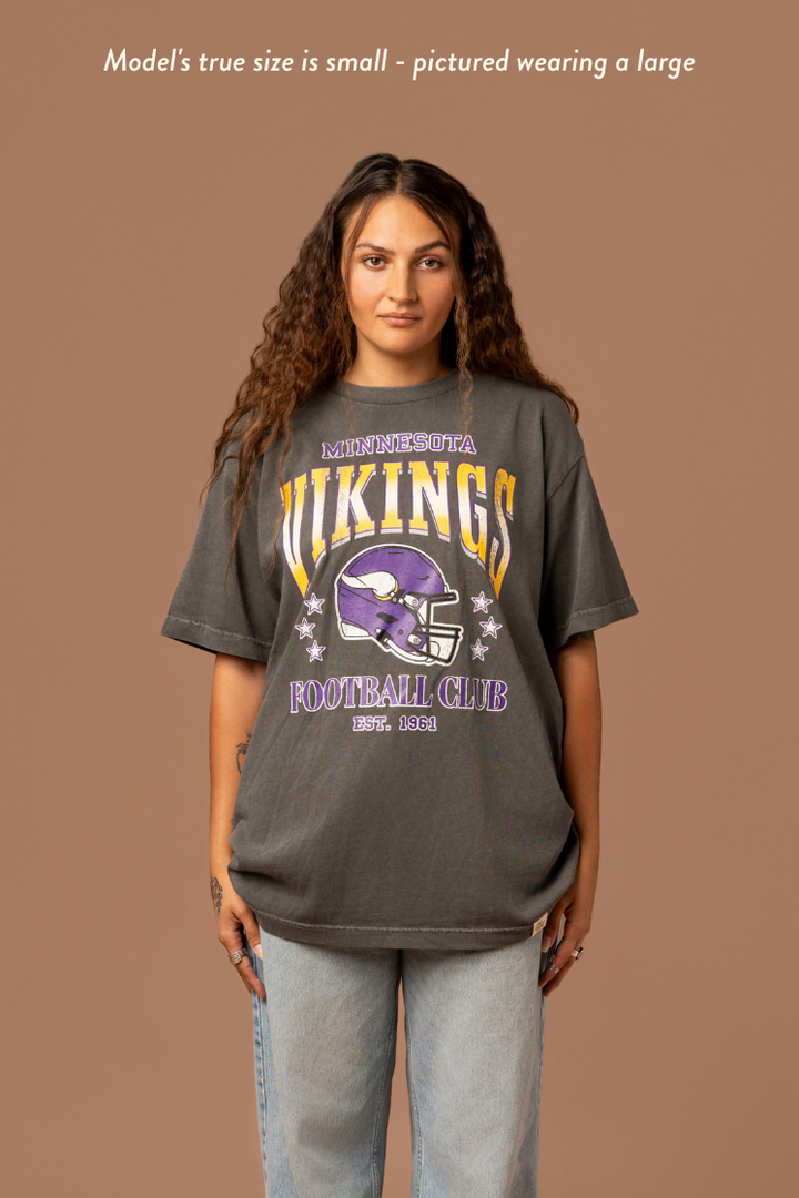 Woman wearing a faded black t-shirt with gold and purple 'Minnesota Vikings Football Club' and 'EST. 1961' text with a purple Vikings helmet graphic in the center. Caption reads "model's true size is small - pictured wearing a large"