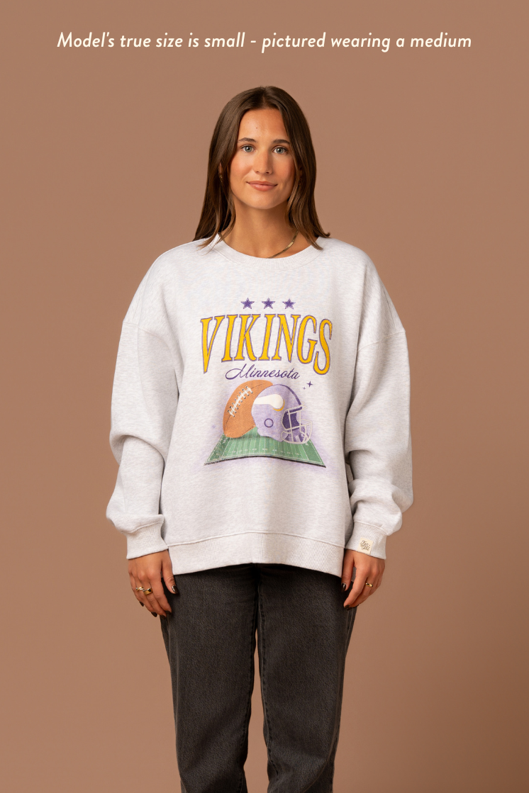 Woman wearing a heather grey crew neck sweatshirt with large yellow 'Vikings' text, purple 'Minnesota' cursive text and a football and helmet graphic under it. Caption reads "Model's true size is small - pictured wearing a medium"
