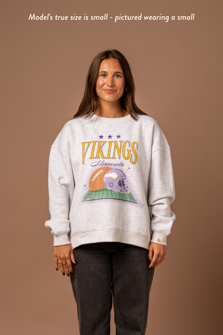 Woman wearing a heather grey crew neck sweatshirt with large yellow 'Vikings' text, purple 'Minnesota' cursive text and a football and helmet graphic under it. Caption reads "Model's true size is small - pictured wearing a small"