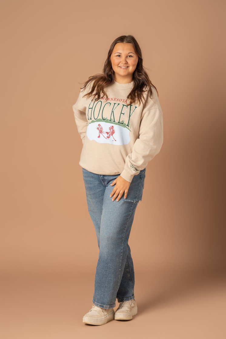 A full body photo of a girl wearing an ivory colored crew neck with a large front design that says Minnesota Hockey with a pond and hockey player graphic.