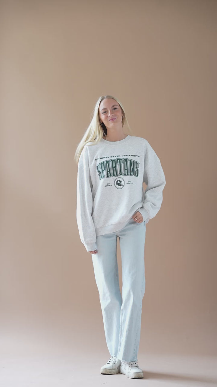 video of a girl wearing a ash gray fleece lined Michigan State University Vintage Oversized Crew Neck sweatshirt with a green, black, and white screenprinted design