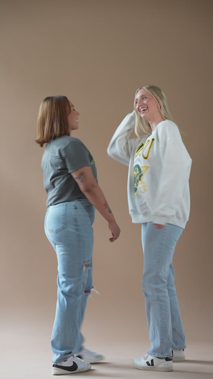 a 13 second video of 2 girls dancing and posing while wearing jeans and one has a black Bison tee on and the other has an ash gray crewneck on with NDSU Football helmet on it in green and gold colors