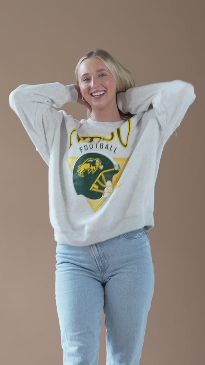 a video of a girl dancing and smiling while wearing a gray crewneck with NDSU football helmet design in green and gold