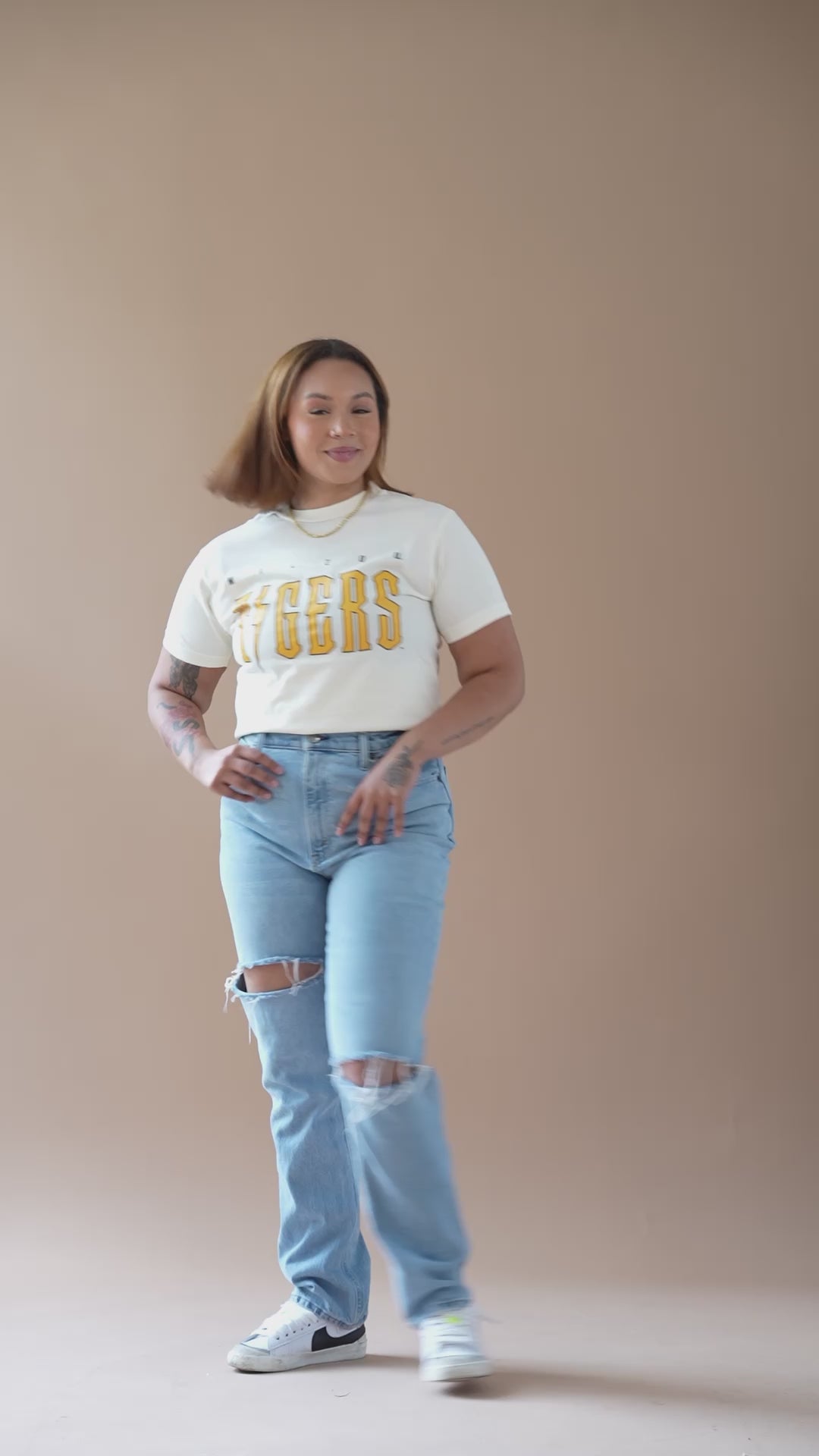 A video of a girl modeling a cream colored t-shirt with a Mizzou Tigers oversized screen print design on the front in black and gold. After a close-up shot of the t-shirt the screen fades to lavender purple and flashes the Fan Girl logo and text "trendy game day apparel."