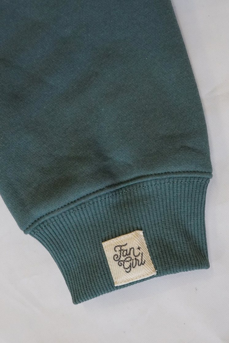 Close-up of a green crew neck sweatshirts sleeve with a branded "Fan Girl" logo