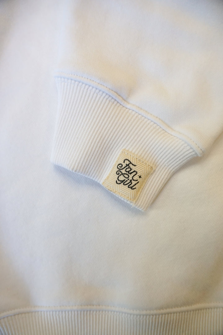 A close up detail photo of the cuff on a white crew neck sweatshirt. The cuff has a small cream patch branded with a Fan Girl logo.