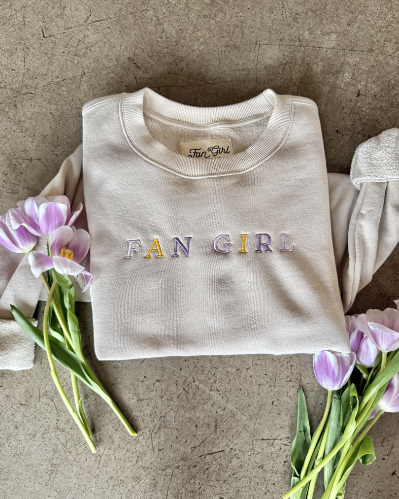 close up picture of a tan crewneck that says Fan Girl and there are flowers laying on the crew